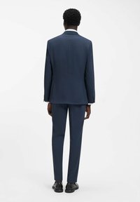 Abito blu navy con finitura testurizzata, caratterizzato da una vestibilità sartoriale, spacco singolo sul retro e scarpe in pelle nere. Colletto di camicia bianca visibile.