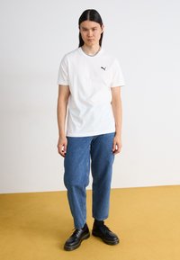 Hvit bomulls T-skjorte med korte ermer og en svart Puma-logo, kombinert med løse blå denimjeans og svarte snøresko.