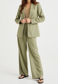 Ljusgrön skräddarsydd blazer och matchande vida byxor, texturerad tyg, öppen front med knytband, kombinerad med en beige topp och svarta flip-flops.