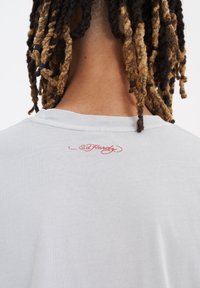 Jasnoszara koszulka z okrągłym dekoltem. Posiada czerwony haftowany logo "Ed Hardy" blisko tylnego kołnierza. Wykonana z miękkiego materiału bawełnianego.