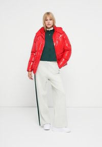 Doudoune rouge brillante avec une capuche, associée à un haut zippé vert foncé et un pantalon gris clair avec une bande latérale verte, ainsi que des chaussures blanches.