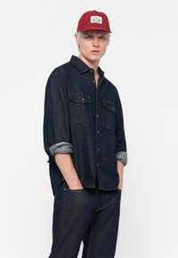 Mørk denimskjorte med knapper foran og to brystlommer, sammen med matchende mørke jeans. Rød caps har et hvitt logo-lapp.