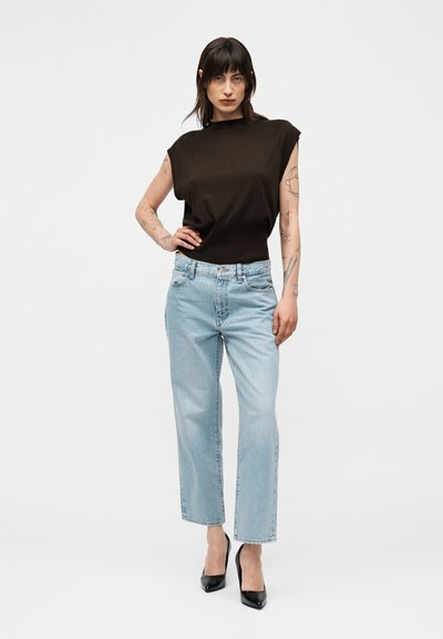 Femme aux cheveux foncés portant un haut noir sans manches, un jean taille haute bleu clair, des talons noirs et plusieurs tatouages visibles sur le bras, debout avec la main sur la hanche.