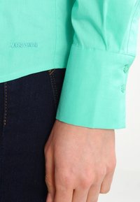 Camicia col verde chiaro con colletto e polsini abbottonati, caratterizzata da una texture liscia e un logo ricamato discreto vicino all'orlo. Jeans scuri visibili sotto.
