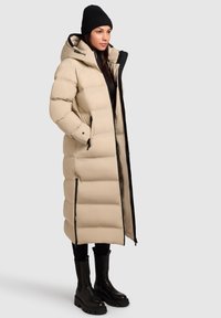 Manteau d'hiver long beige avec capuche, accents noirs, fermetures éclair et poches latérales. Associé à des bottes noires et un bonnet en tricot.