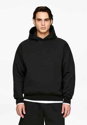 Młody mężczyzna stoi, ubrany w prostą czarną bluzę z kapturem i sznurkami, biała koszulka widoczna u dołu oraz czarne spodnie.