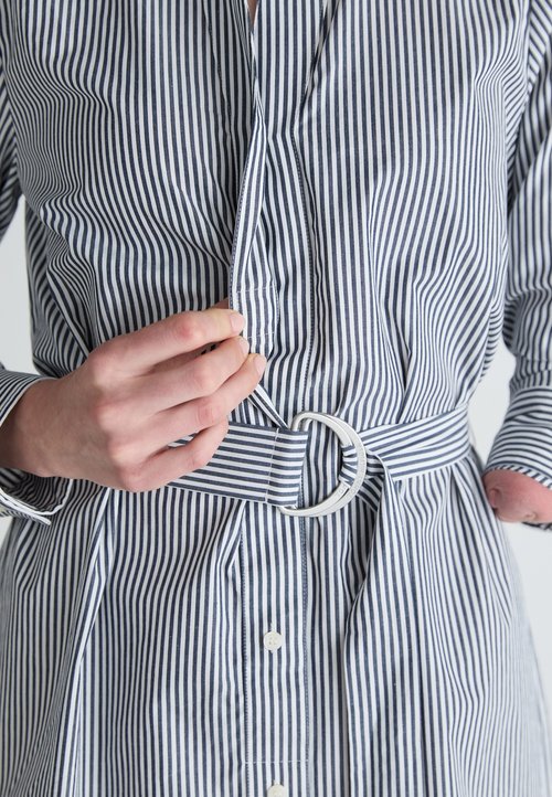Stripet skjorte med blå og hvite vertikale striper, knappestolpe foran og belte i midjen med en rund metallspenne. Teksturen er glatt.