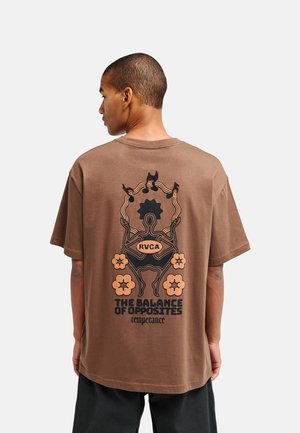Man draagt een bruin T-shirt met een abstracte menselijke figuur, bloemen en de tekst "The Balance of Opposites temperance" op de achterkant.