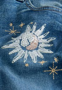 Denimová látka s motivy slunce a hvězd ve stříbrných flitrech a béžových stehách na modrém pozadí, s texturovaným povrchem.