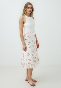 Weißer Spitzenrock mit floralen Mustern in Pink und Grün, kombiniert mit einem eng anliegenden weißen Tanktop. Das Model steht in entspannter Pose.