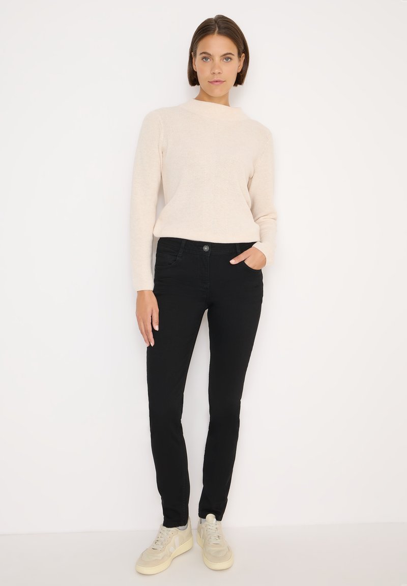 Jeans skinny neri abbinati a un maglione a maniche lunghe color beige chiaro. Il maglione presenta un collo alto e una texture morbida.