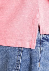 Rosa bomullströja med korta ärmar, med en sidospalt nederkant och en texturerad yta, lagd över blå denimjeans.