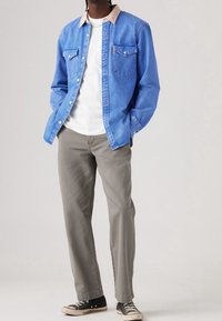 Person wearing blue denim jacket over white shirt, gray pants, and black sneakers standing with hands in pockets against plain background.