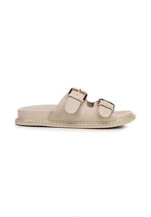 Sandalia plana de ante beige con dos tiras ajustables que cuentan con hebillas en tono dorado y una suela texturizada plana.