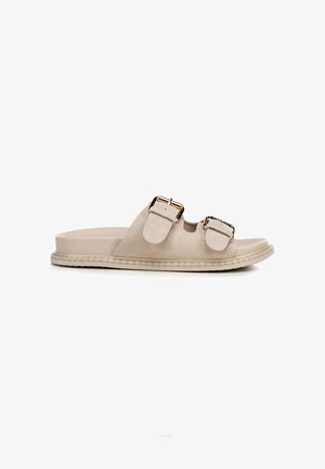 Sandalia plana de ante beige con dos tiras ajustables que cuentan con hebillas en tono dorado y una suela texturizada plana.