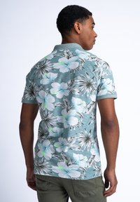 Puuvartinen, lyhythihainen polo teal-värissä, jossa on kukkakuosi valkoisten ja vihreiden kukkien kera. Materiaali on kevyttä ja teksturoitua.