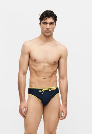 Paita pois mies, jolla on lyhyet tummat hiukset, pukeutunut tummansinisiin uimahousuihin, joissa on keltaiset reunukset ja etupuolella keltainen nyöritys, seisoo yksivärisen taustan edessä.