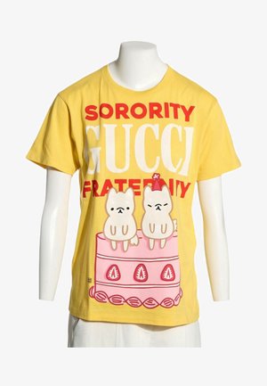 Gelbes T-Shirt mit dem Text "Sorority Gucci Fraternity" und einer Illustration von zwei kleinen Hunden, die auf einem rosa Kuchen mit Erdbeer-Dekoration sitzen.