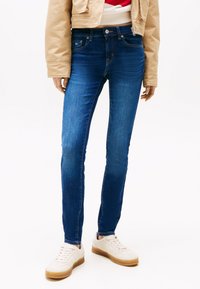 Mörkblå slim-fit jeans i denim med midjehöjd och fem fickor, kombinerade med vita sneakers och en beige kort jacka.