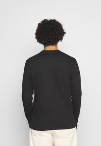 Černá dlouhá rukávová košile s kulatým výstřihem, vyrobená z hladké tkaniny, zobrazená ze zadní strany, v kombinaci se světle barevnými kalhotami.