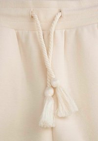 Beżowy sznurek ściągający z dwoma chwostami, wykonany z skręconego sznura, nad teksturowanym materiałem w dopasowanym kolorze. Widoczny szczegół miękkiej, gładkiej powierzchni.