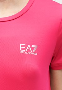 Κοντινό πλάνο σε ροζ μπλουζάκι με λευκό λογότυπο "EA7 Emporio Armani" στο πάνω μέρος του στήθους.