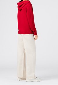 Czerwony sweatshirt z kapturem i długimi rękawami, zestawiony z szerokimi, kremowymi spodniami. Spodnie mają postrzępiony dół, noszone z jasnymi, sportowymi butami.