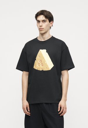 Νέος άνδρας που φοράει μαύρο υπερμεγέθες μπλουζάκι με μεγάλο κίτρινο γραφικό ελβετικού τυριού μπροστά, στέκεται σε απλό λευκό φόντο.