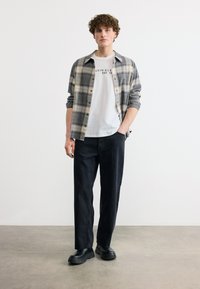 Νεαρός άνδρας με σγουρά μαλλιά που φοράει λευκό t-shirt Calvin Klein, γκρι καρό πουκάμισο, μαύρο παντελόνι και μαύρα παπούτσια, στέκεται μπροστά σε ένα απλό φόντο.