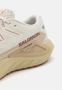 Helle-beige Laufschuh mit Mesh-Obermaterial, verstärkter Ferse, braunen Akzenten und strukturiertem Gummisohle mit dynamischem Profil.
