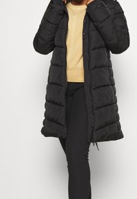 Abrigo acolchado negro con un diseño largo y ajustado, que presenta un cierre de cremallera, bolsillos laterales y un patrón de acolchado texturizado. Lleva puesto un top beige.