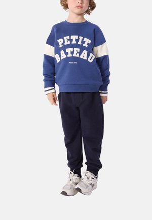 Sweatshirt bleu avec le texte blanc "PETIT BATEAU" et des poignets rayés, associé à un pantalon de survêtement bleu marine et des baskets blanches.