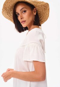 Witte blouse met korte mouwen, een gather in de taille, kanten details en een losse pasvorm. Gecombineerd met een grote strohoed en ringoorbellen.