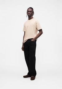 Beżowy bawełniany t-shirt z krótkimi rękawami i małym niebieskim logo, w połączeniu z czarnymi dżinsami, noszony z brązowymi zamszowymi butami.