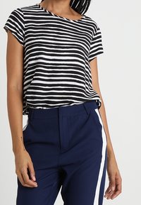 Mujer vistiendo una blusa de rayas horizontales en blanco y negro de manga corta, metida dentro de un pantalón azul marino con una franja vertical blanca a los lados.