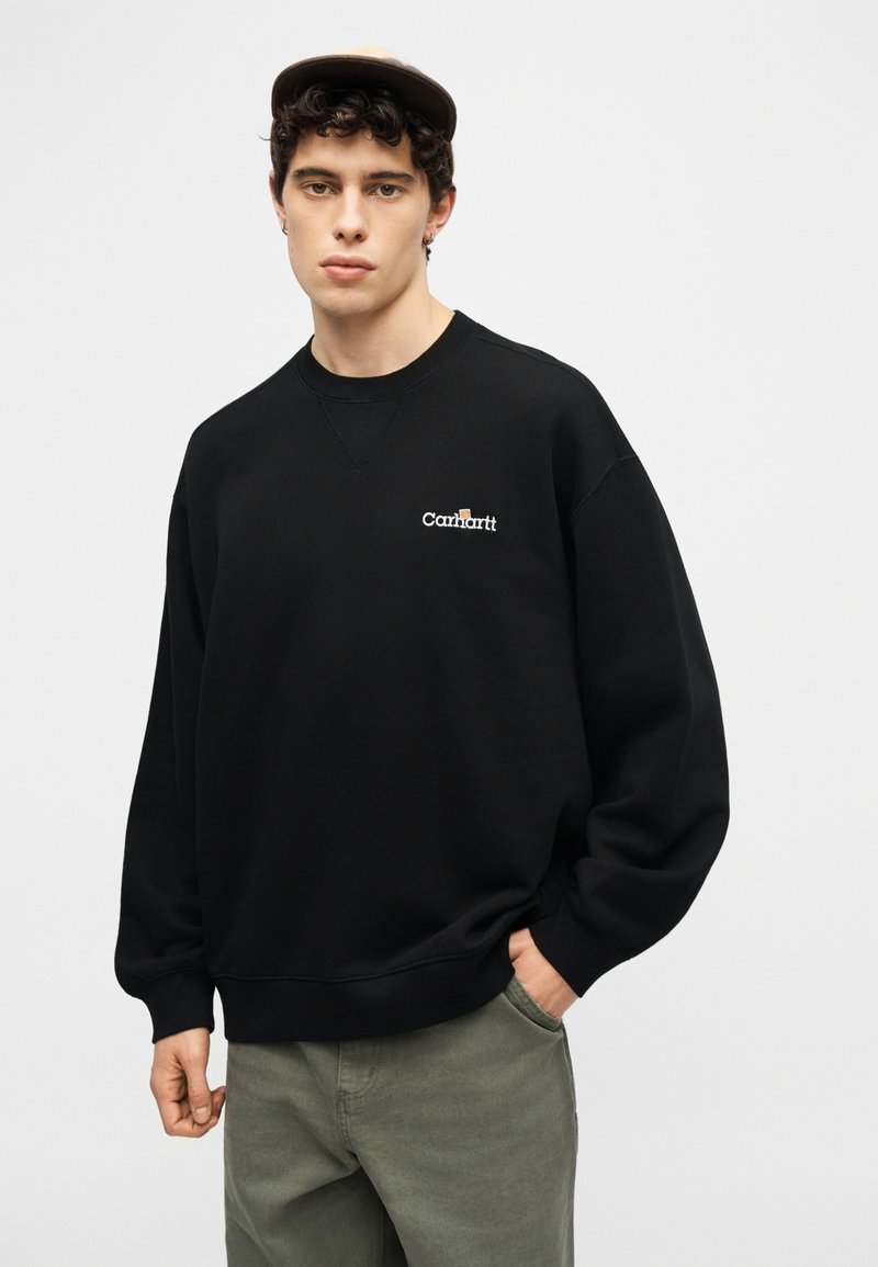 Giovane uomo con indosso una felpa Carhartt nera, pantaloni verdi e un cappello beige, in piedi con una mano in tasca su uno sfondo semplice.