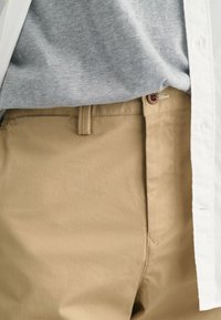 Khaki pants made of cotton blend with a flat front, button closure, and side pockets; paired with a gray t-shirt underneath a white shirt.