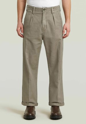 Mann trägt hochgekrempelte beige plissierte Hose und braune Lederstiefel, steht vor einem einfarbigen hellen Hintergrund.