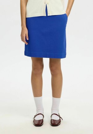 Žena v modrej sukni po kolená, biele ponožky a hnedé Mary Jane topánky, stojaca s jednou rukou v vrecku na bielom pozadí.