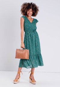 Green patterned dress with a V-neckline and ruffled sleeves. Brown leather handbag and tan strappy wedge sandals.