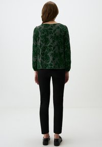 Blusa verde con motivo floreale, a maniche lunghe, in tessuto trasparente e con polsini arricciati, abbinata a pantaloni slim fit neri e scarpe nere.