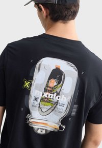 Černé tričko s potiskem skleněné nádoby. Uvnitř se nachází bota s oranžovými akcenty a zelenými detaily, pod designem je text a logo.