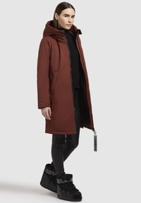 Cappotto lungo rosso impermeabile con cappuccio, chiusura con zip, tasche laterali, abbinato a leggins in pelle nera e stivali alti neri.