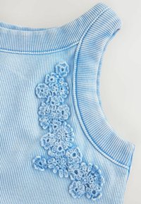 Helles blaues ärmelloses Oberteil mit gerippter Textur, das auf der linken Seite ein dekoratives gehäkeltes Blumenmuster aufweist.