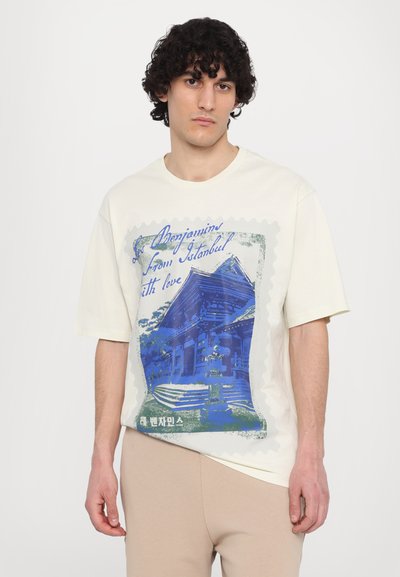Cremfarbene T-Shirt mit einem blauen Grafikdruck eines Gebäudes, umrahmt von einem stempelartigen Rand, mit Text in Schreibschrift und Blockbuchstaben.