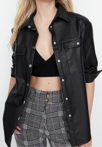 Giacca camicia in ecopelle nera con bottoni a pressione, due tasche sul petto e manicotti arrotolati, abbinata a pantaloni a quadri a vita alta.