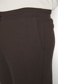 Brown cotton jogger pants feature a soft texture, elastic waistband, and side pockets, shown from the waist down in close-up.