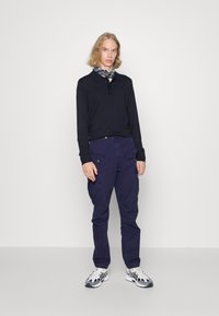 Polo a maniche lunghe blu navy con colletto, abbinato a pantaloni cargo scuri con tasche laterali e una sciarpa con stampa classica. Scarpe da ginnastica.