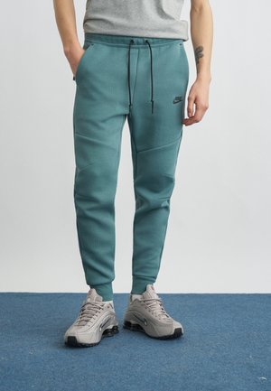 Osoba ubrana w turkusowe dresy Nike z czarnymi sznurkami i beżowymi butami Nike stoi na niebieskim dywanie.