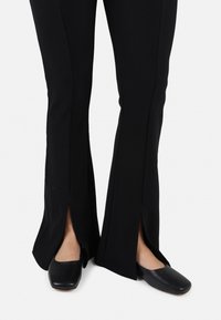 Black flared pants with side slits, made from textured fabric, paired with black closed-toe shoes featuring a smooth finish and low heel.